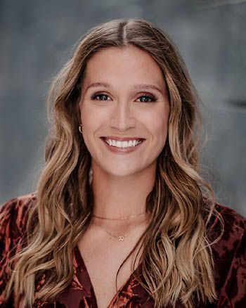 Woman with long wavy blonde hair smiling at the camera, wearing a dark floral top.