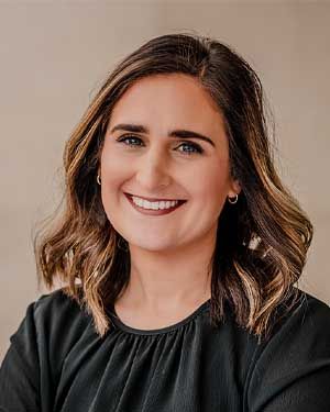 Woman with brown hair smiles, wearing a black shirt, in front of a neutral background.