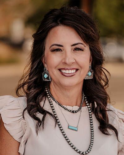 Woman smiling, wearing a beige blouse, turquoise jewelry, and a beaded necklace; outdoors.