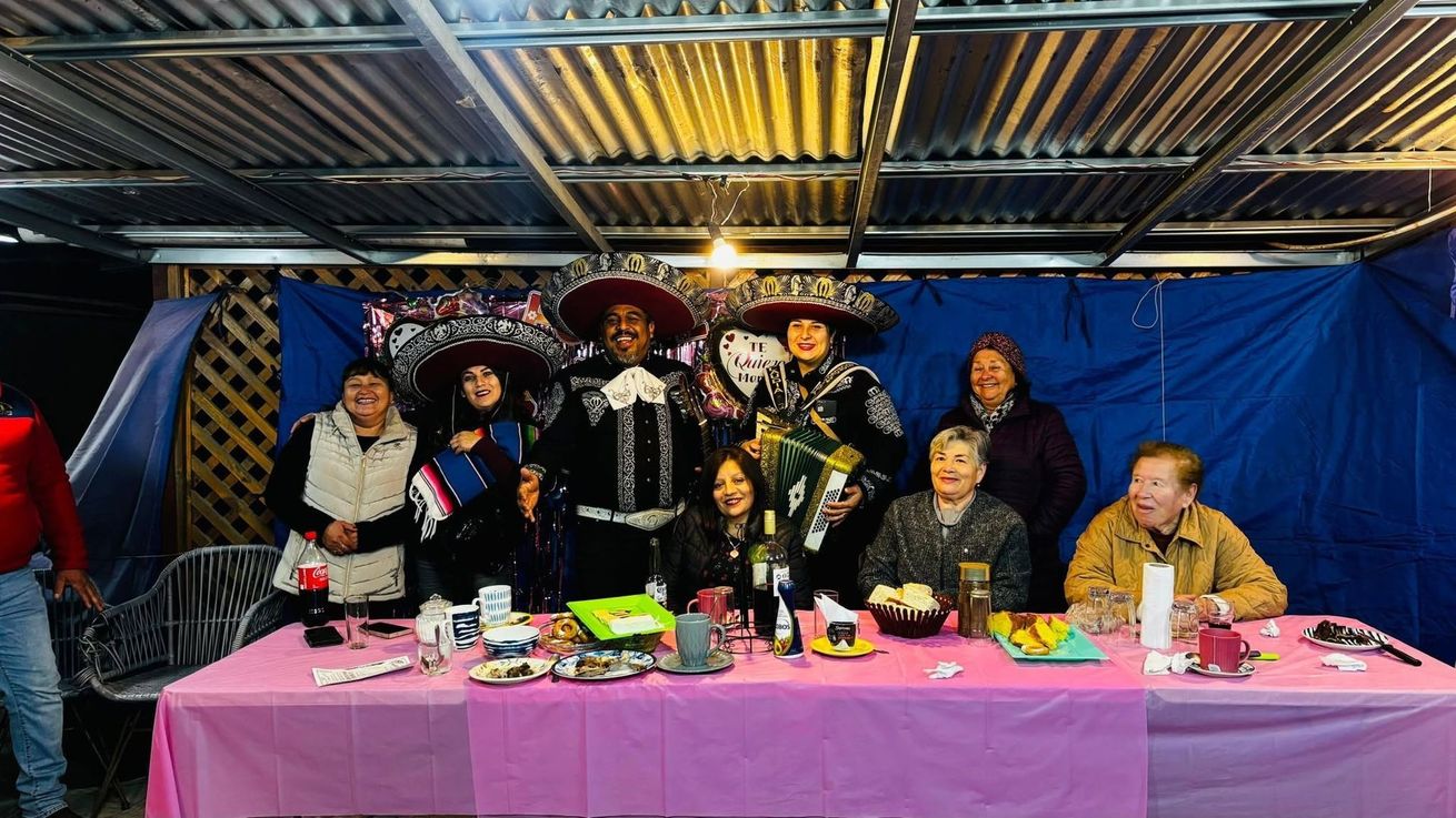 Grupo de personas disfrazadas posando detrás de una mesa rosa bajo un techo de chapa ondulada con fondo azul.