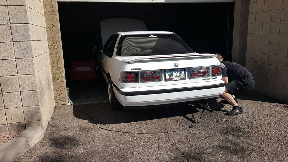 White sedan in a garage while a person kneels beside the rear tire, using a jack or tool.