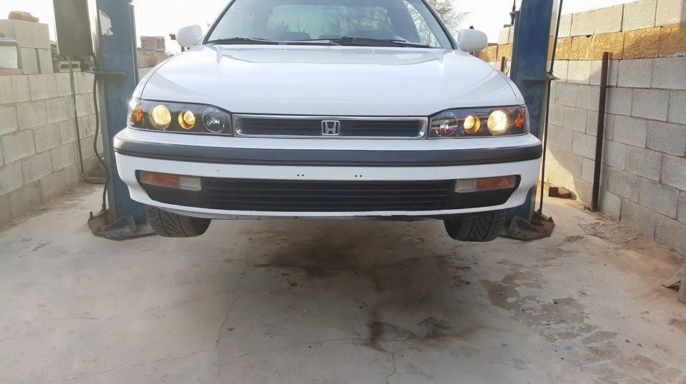 White sedan parked between cinder block walls, front view with headlights on.