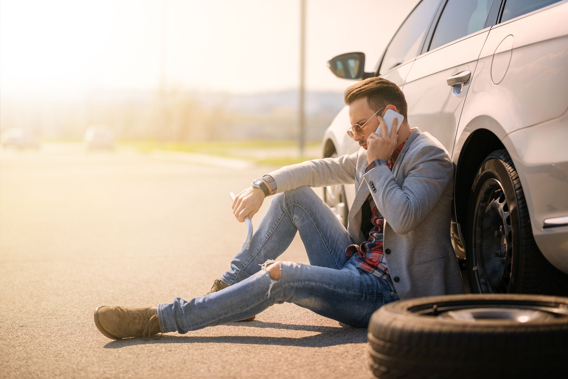 A man is sitting on the ground next to a car talking on a cell phone.