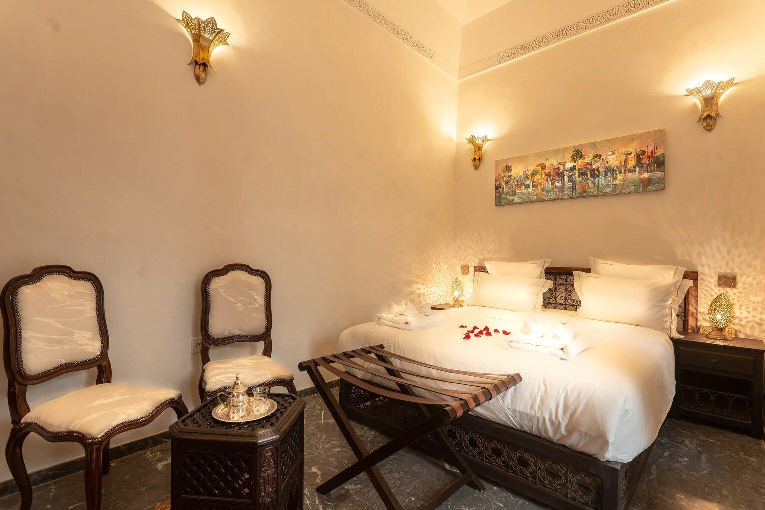 Bedroom with ornate furniture, bed with rose petals, two chairs, small table, and decorative wall sconces.
