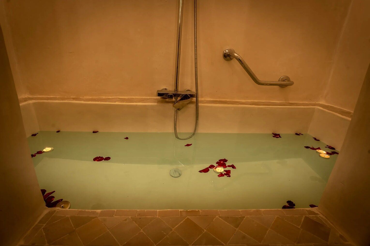 Bathtub filled with water and rose petals, with a showerhead and grab bar in a warm-toned bathroom.