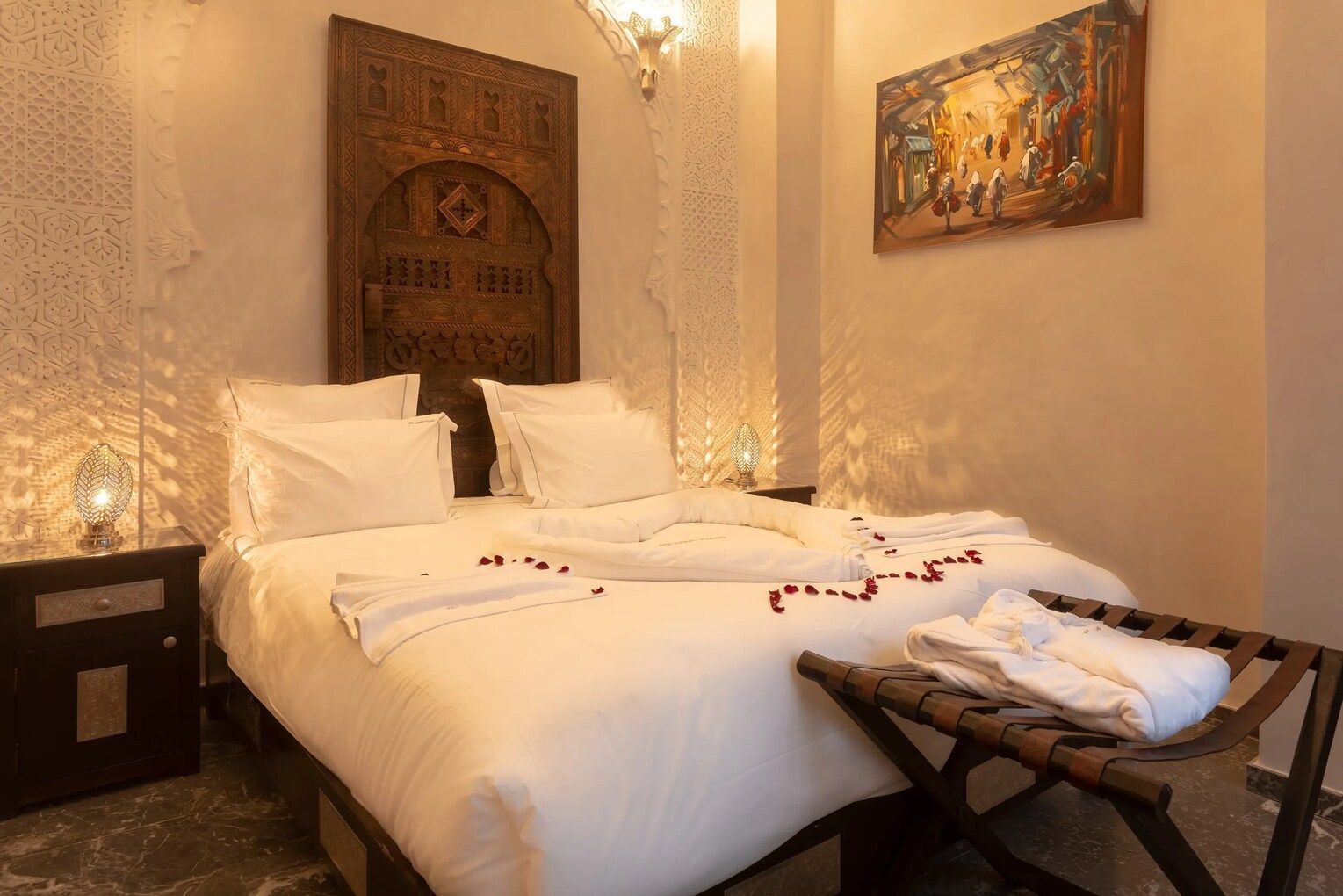 Cozy bedroom with a double bed. Features ornate headboard, nightstand, and art. Robe and rose petals on bed.