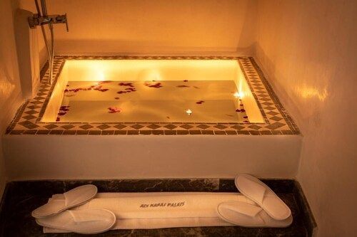 Bathtub filled with water and rose petals, lit by soft lights, with slippers and a towel laid out.