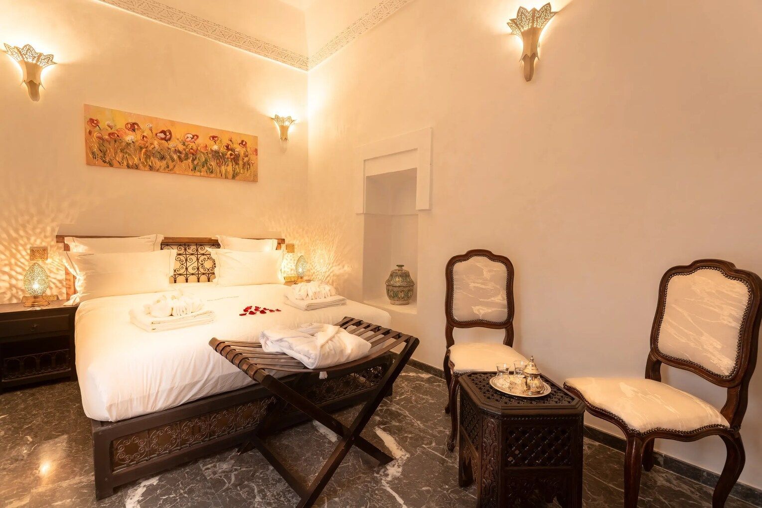 Cozy hotel room with ornate carved bed, chairs, and bedside tables; white walls; soft lighting.