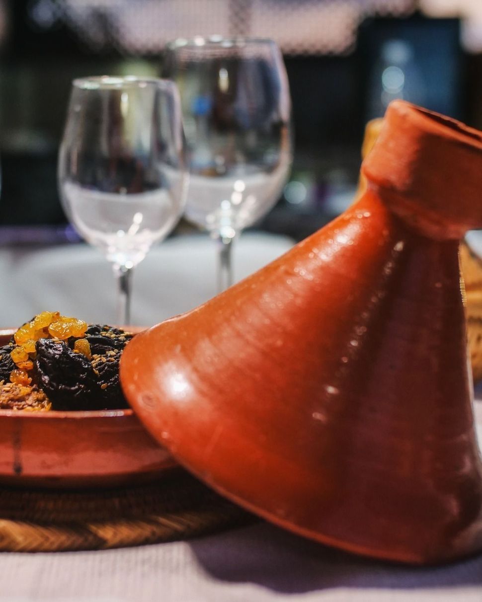 A reddish-brown tagine dish with the conical lid askew, filled with food; wine glasses in background.