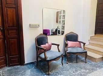 Two antique chairs with pink pillows under a rectangular mirror on a wall; stairs to the right.