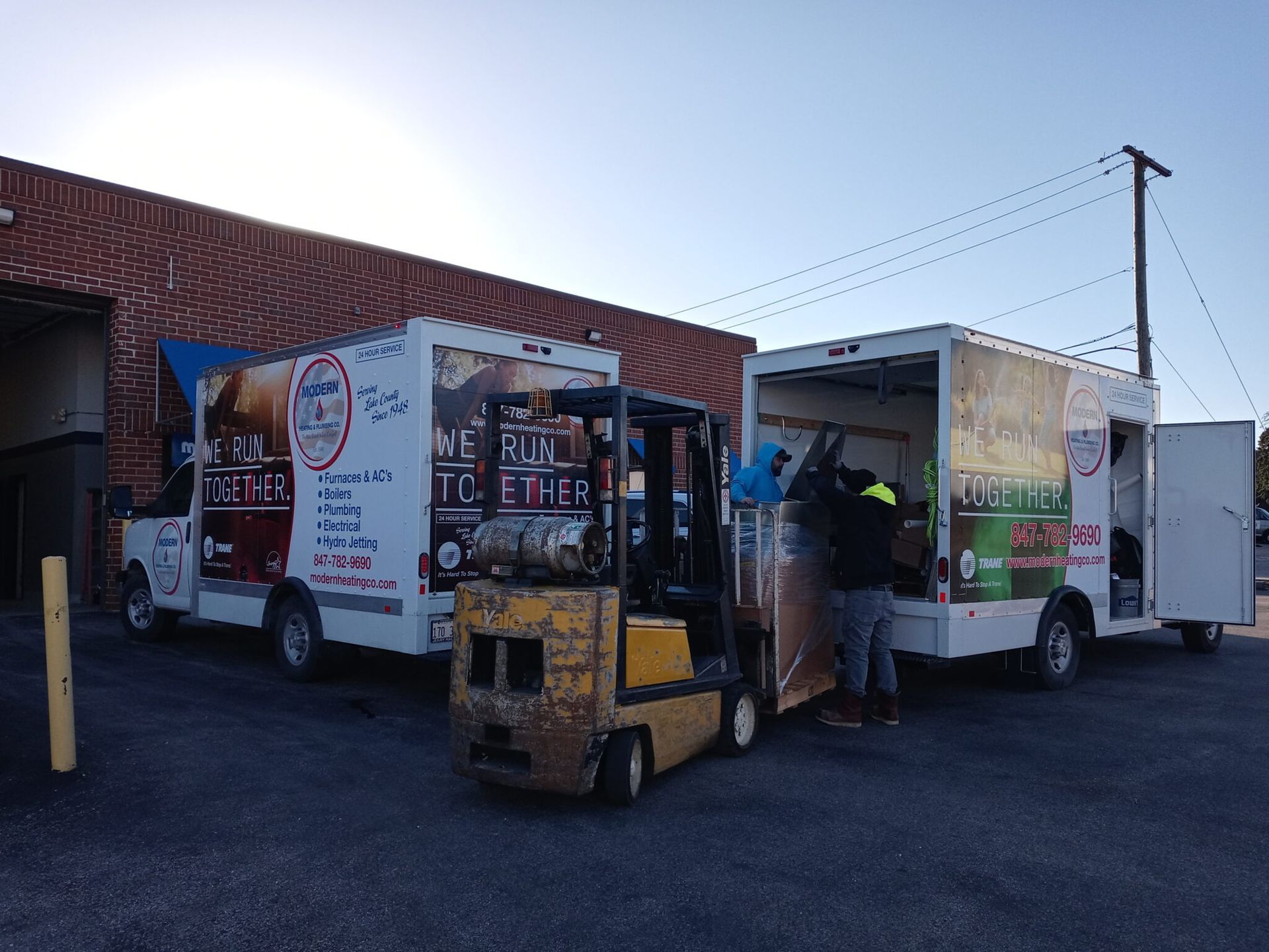 Forklift loading a truck next to another truck; worker in doorway; brick building and utility pole in background.
