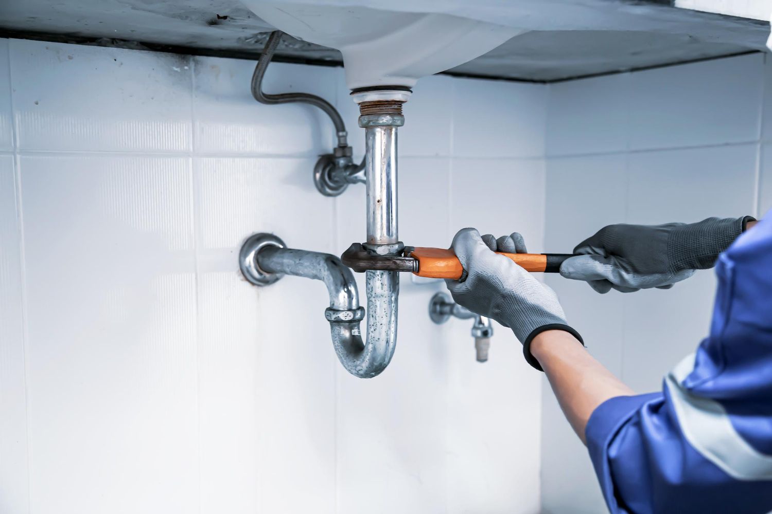 Plumber in blue uniform uses a wrench to tighten pipes under a sink.