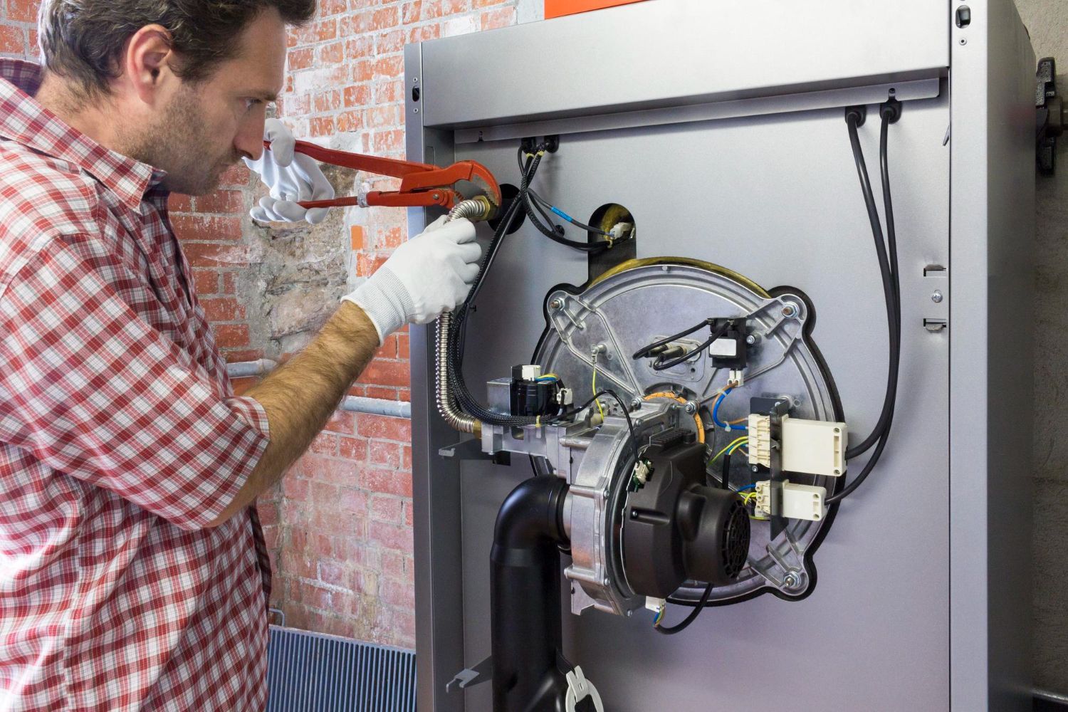 A person in a red plaid shirt and gloves repairs a furnace with pliers.