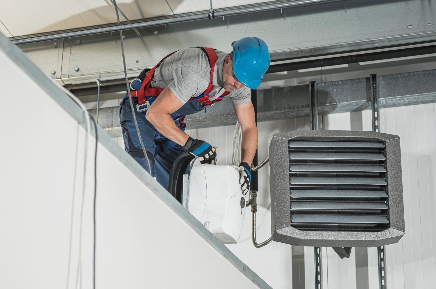HVAC technician in blue hard hat works on a large heater, high up in a warehouse.