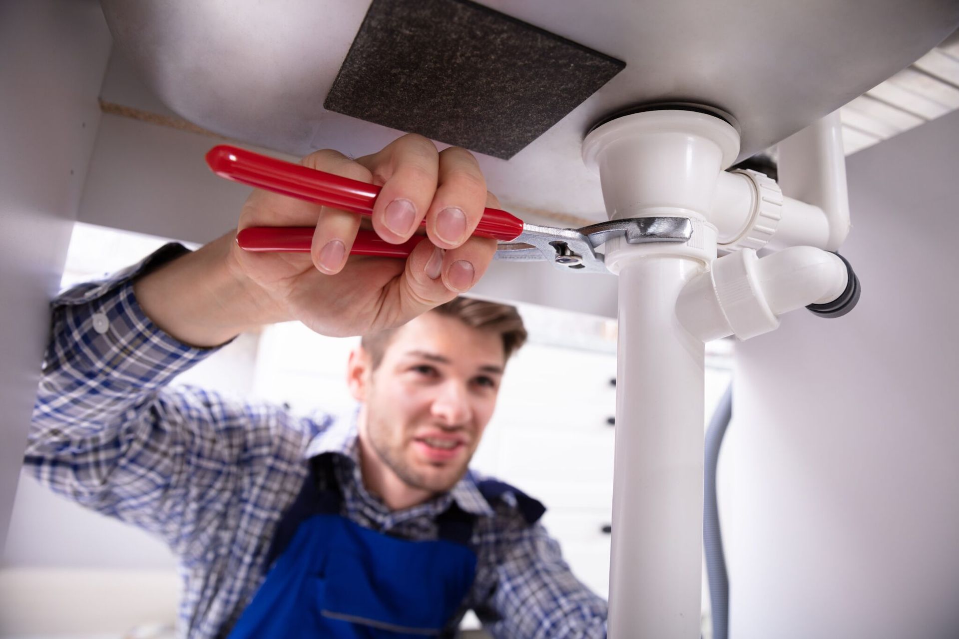 Plumber using a red wrench under a sink, working on white pipes.
