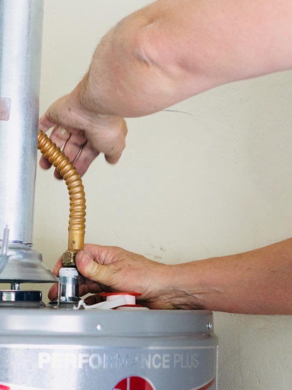 Hands connecting a flexible gas line to a water heater. The heater is silver with
