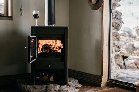 Black wood-burning stove with open door, flames visible. Wine glass on top. Inside rustic room with open doorway.