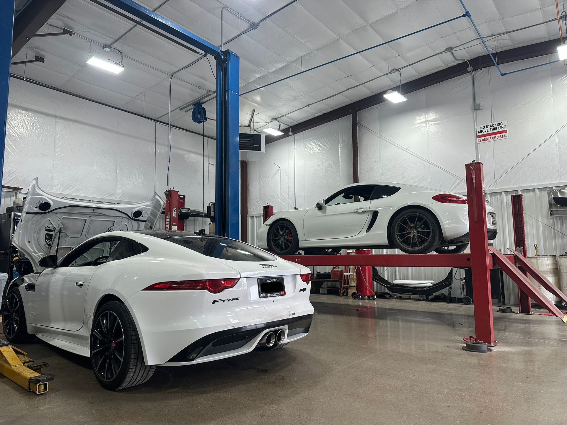 Two white sports cars in a bright auto shop on lifts, with tools and a blue service post.