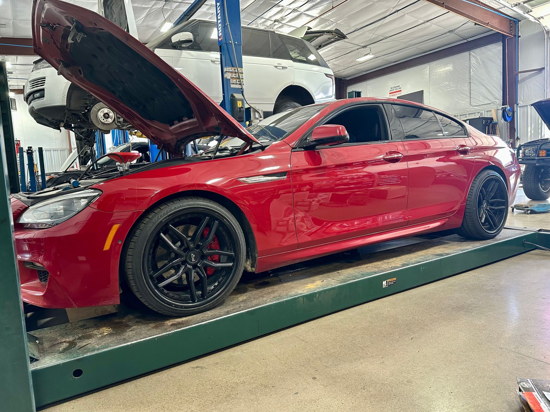 Red sports sedan on a lift in an auto shop, hood open, with a white car raised in the background
