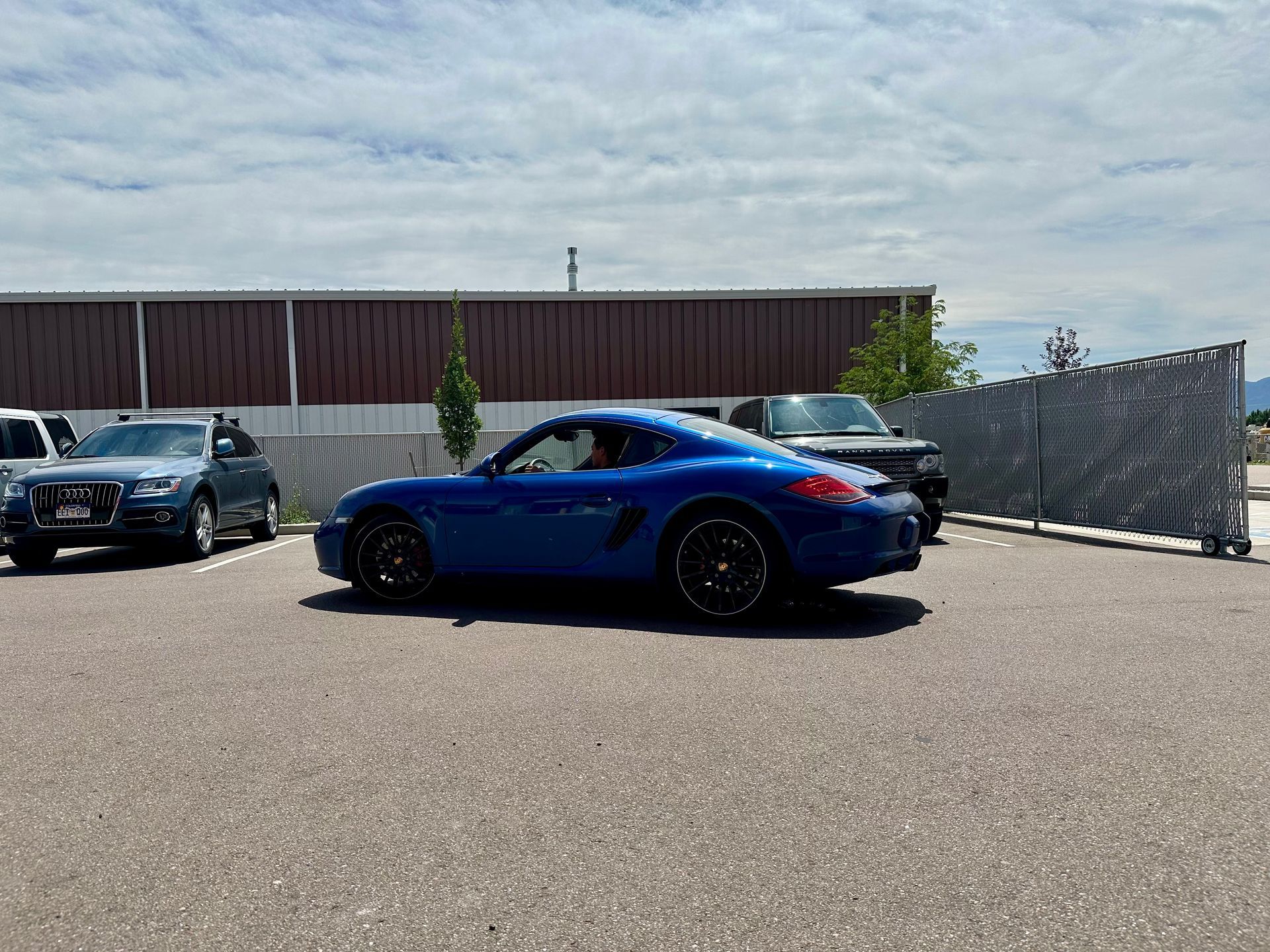 Blue sports car parked in a lot beside other cars and a fence under a cloudy sky