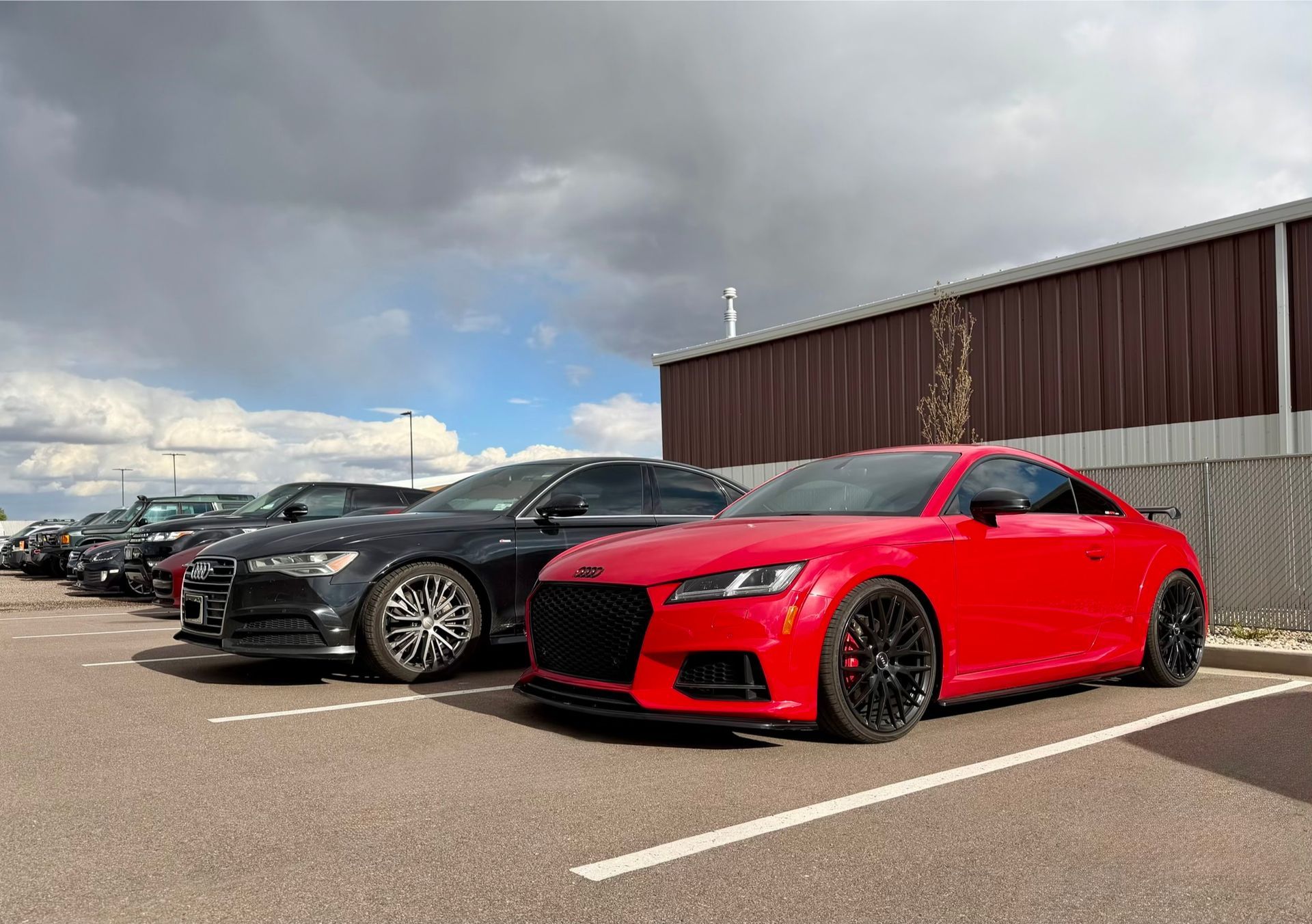 Red sports coupe parked in front of a line of cars in a parking lot under cloudy skies