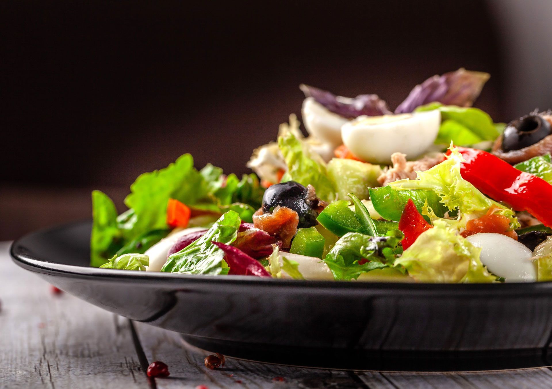 Salad in black bowl with lettuce, olives, egg, and peppers on a white wooden surface.