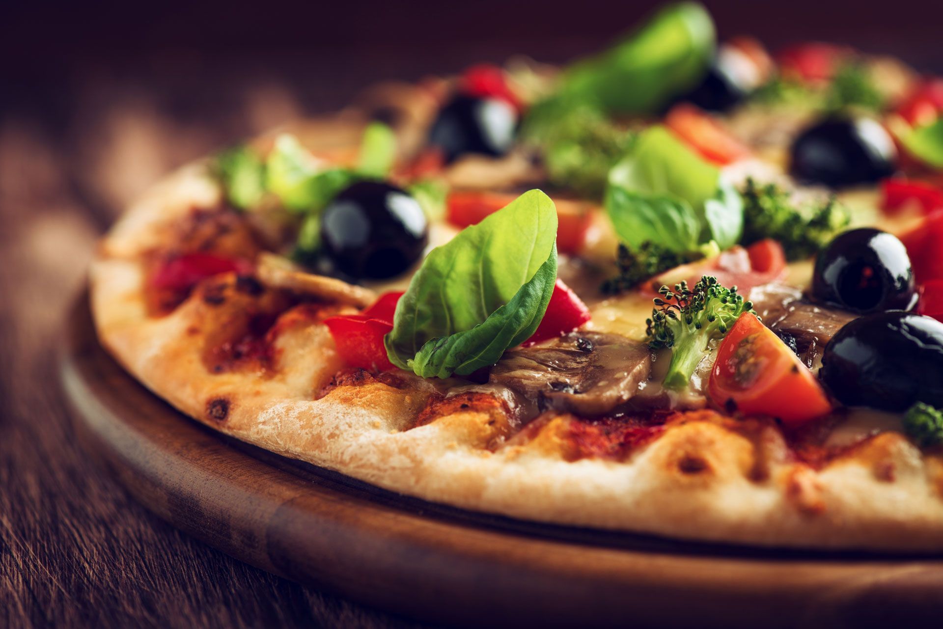 Pizza with black olives, red peppers, tomatoes, and basil on wooden board.