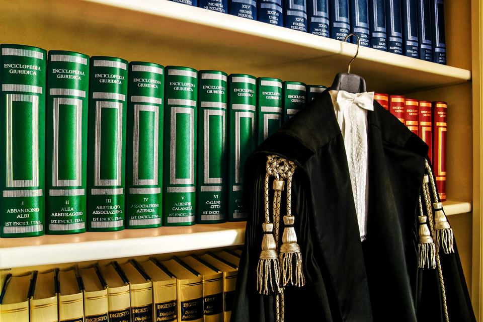 Libreria con libri di giurisprudenza con il dorso verde e una toga nera da giudice con colletto bianco e nappe.