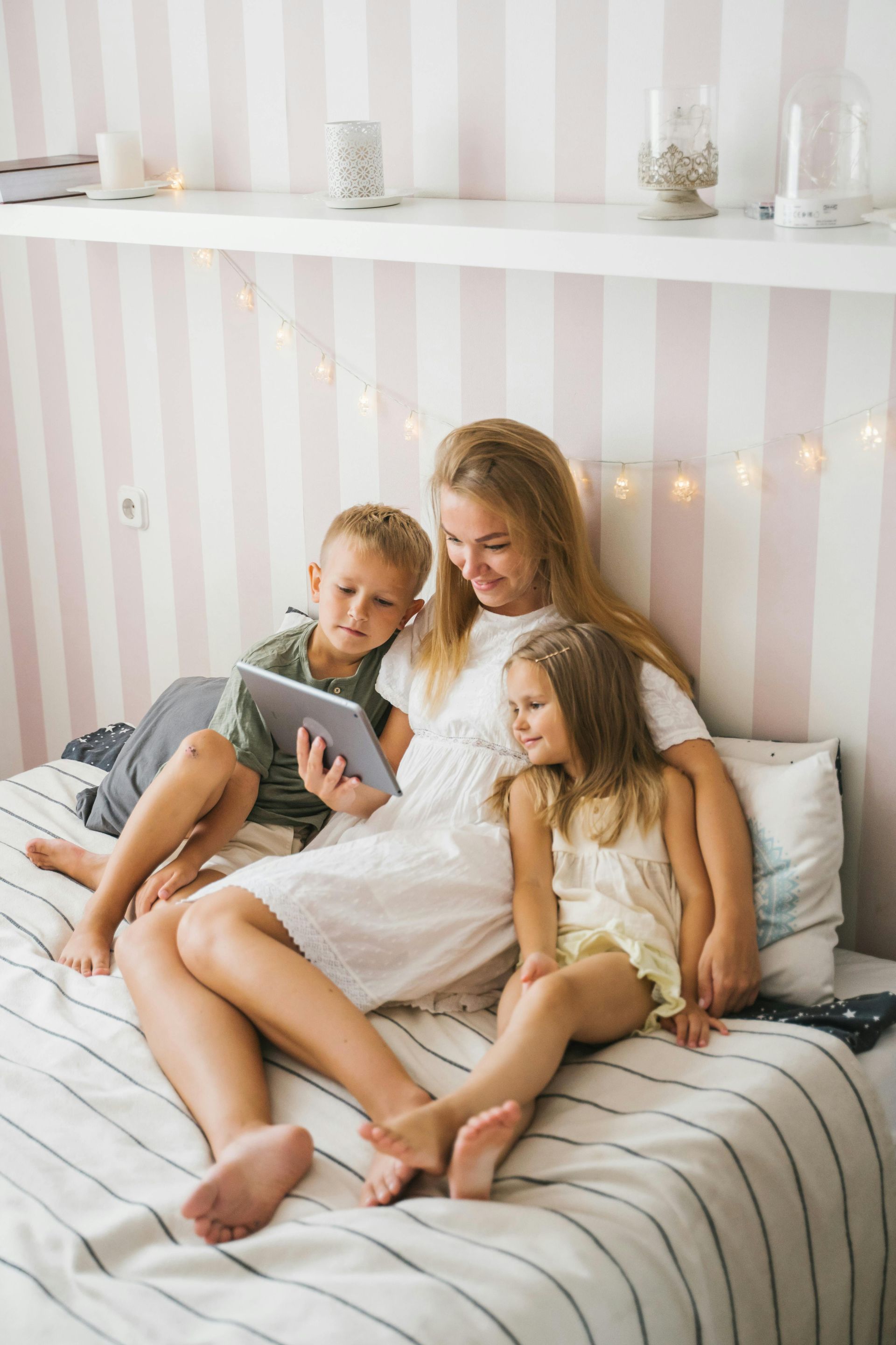 Donna e due bambini su un letto, che guardano un tablet. Parete a strisce bianche e rosa chiaro, lucine.