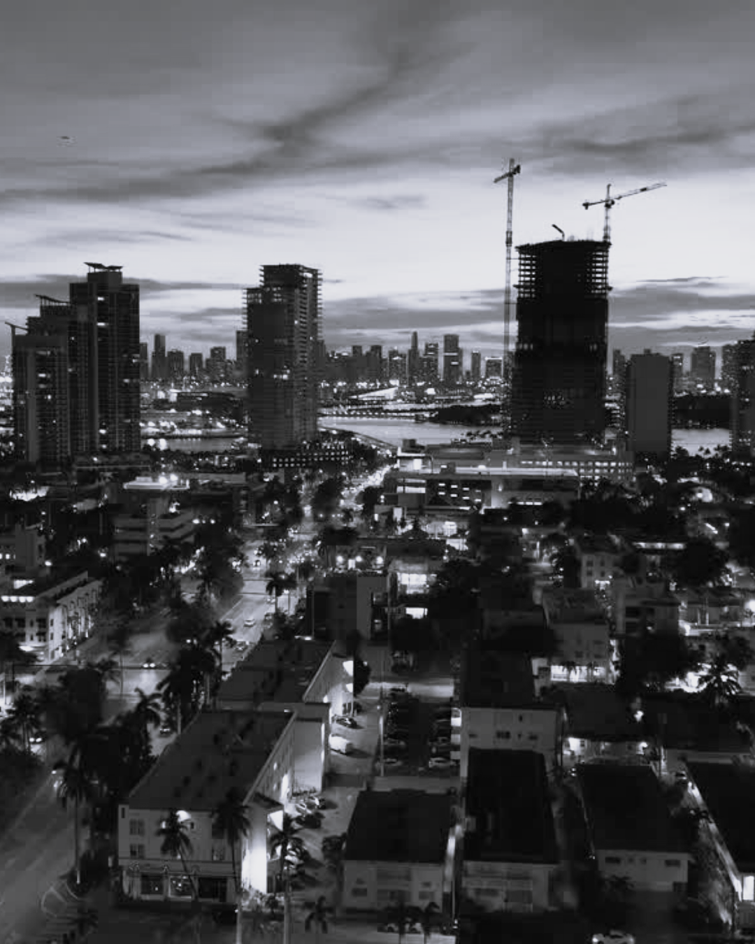 Black-and-white city skyline at dusk with high-rise buildings, cranes, and lit streets of Miami FL