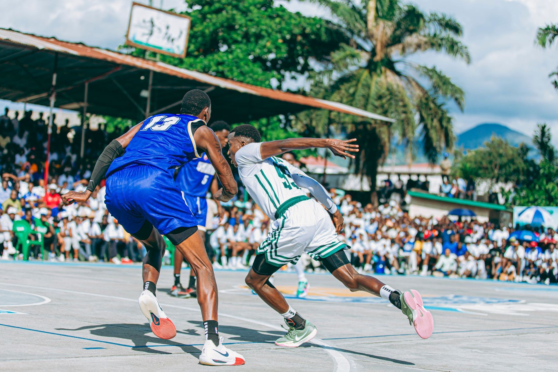 Two basketball players in blue and white jerseys dribble on an outdoor court with spectators nearby