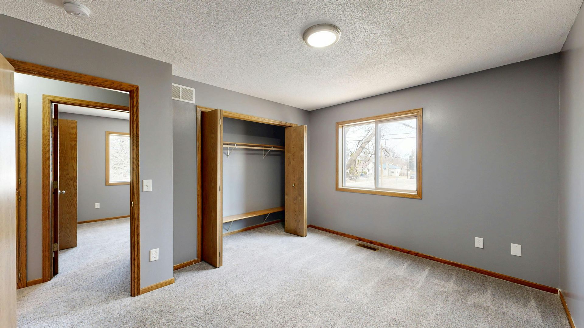 Empty beige bedroom with carpet, open closet and doorway, and a window letting in daylight.