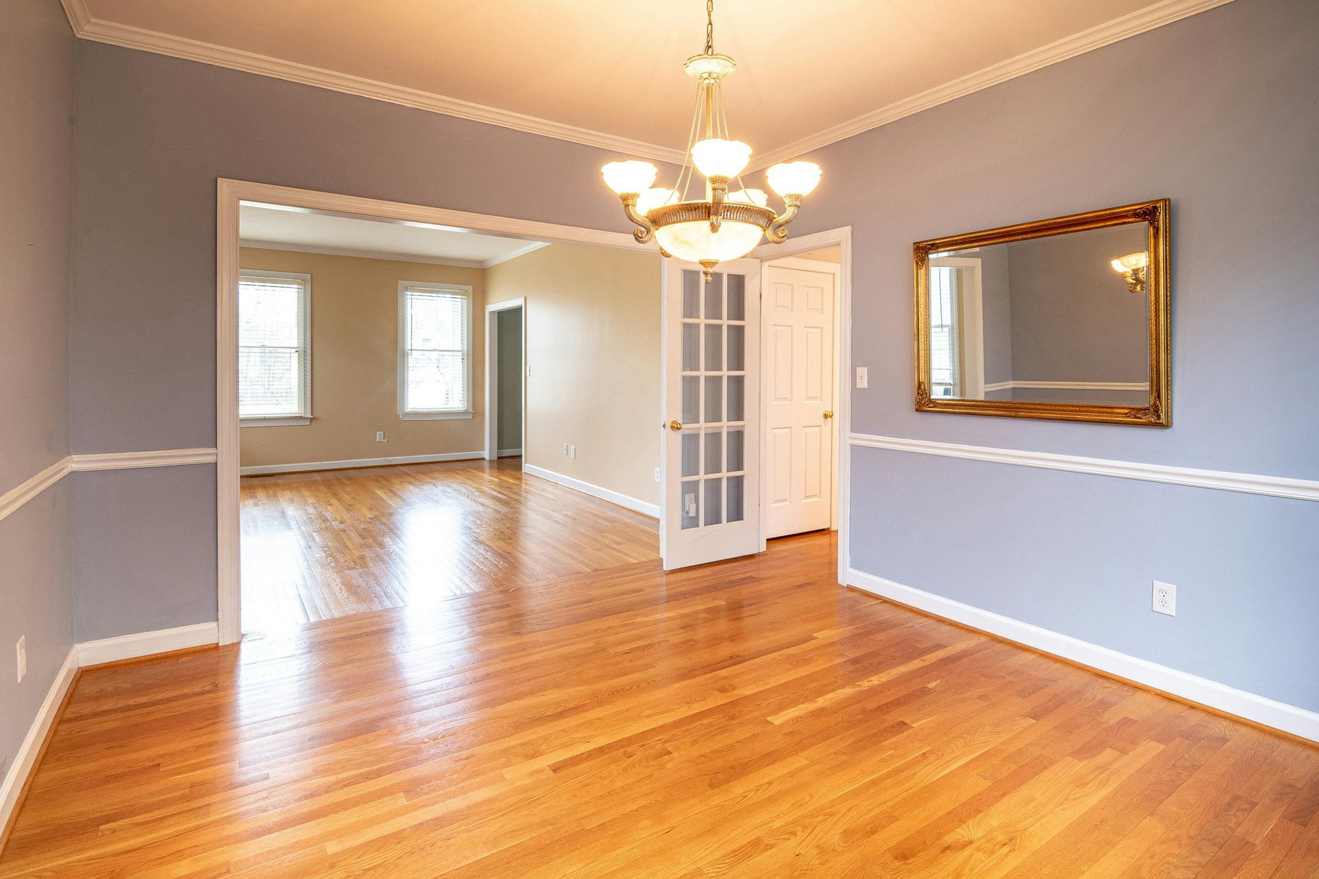 Empty dining room with hardwood floors, gray walls, white trim, chandelier, and a large mirror on the wall