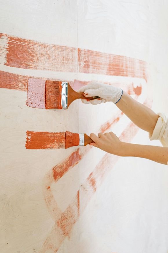 Two hands painting wide orange strokes on a white wall with a brush and roller