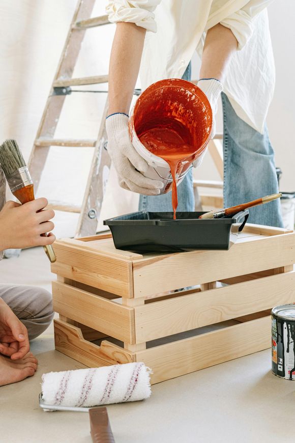 Person pours red paint into a tray while painting supplies and a wooden crate sit on the floor.
