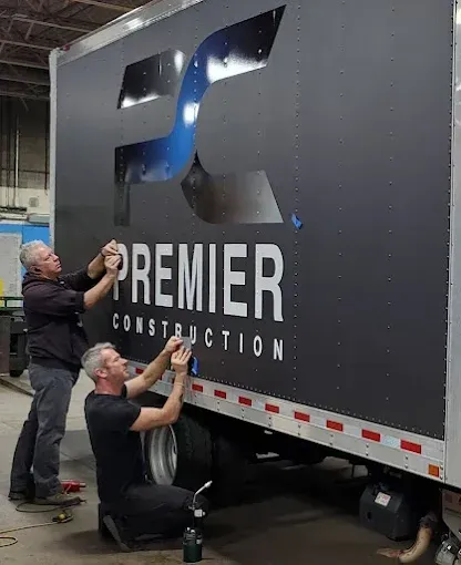 Two workers applying a large blue logo decal to a black Premier Construction truck trailer.