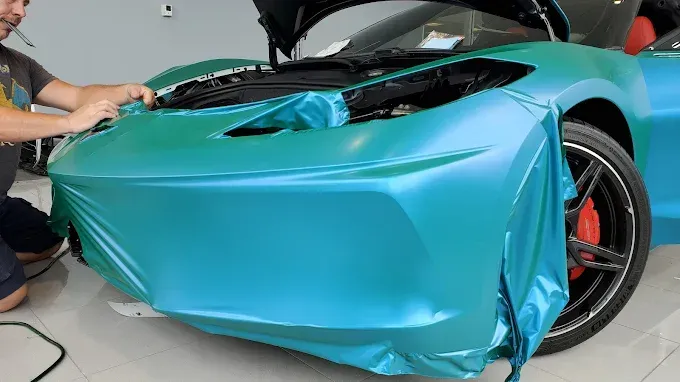 Person wrapping a turquoise sports car in blue vinyl inside a garage.