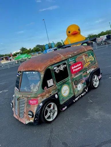 Rusty vintage van with colorful decals and a giant yellow duck on top in a parking lot