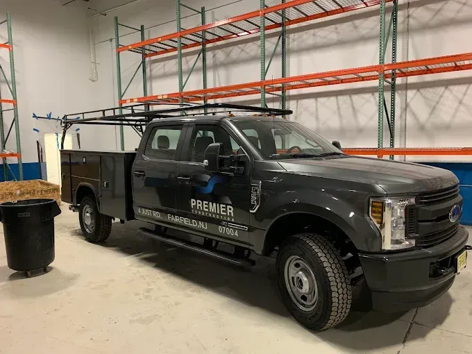 Black utility truck with ladder rack parked inside a warehouse beside metal shelving racks.
