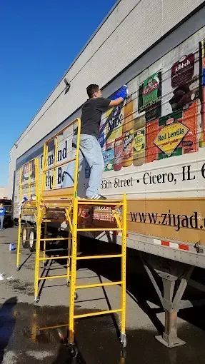Person on yellow ladder painting a large mural on the side of a truck trailer.