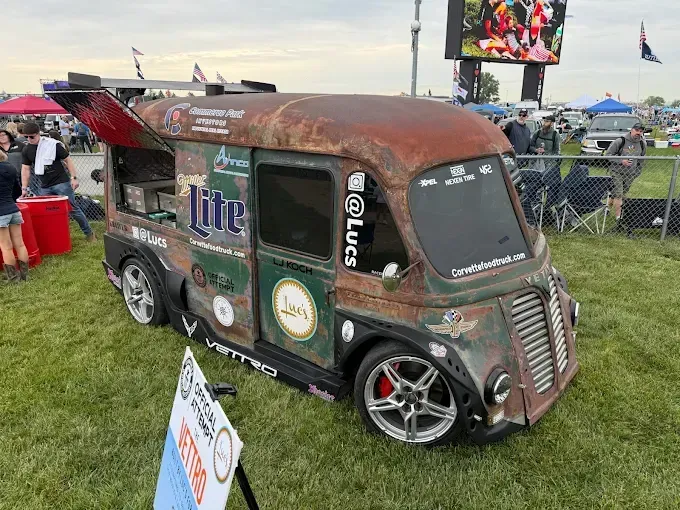 Rusty custom van with beer decals at an outdoor event, parked on grass near a crowd and screens.