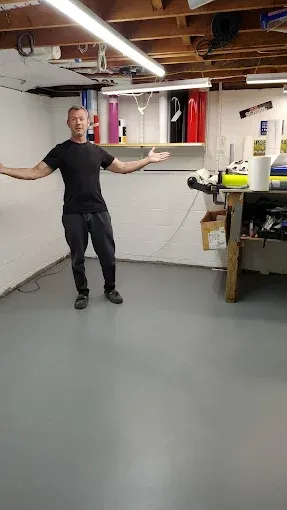 Person in black shirt stands in a basement workshop, gesturing toward a workbench and shelves.