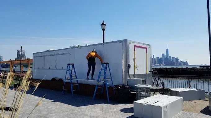 Two workers on ladders repairing a white rooftop structure with a city skyline in the background