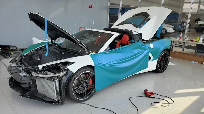 Blue-and-black sports car in a workshop, hood and trunk open, with a person working on the front end.