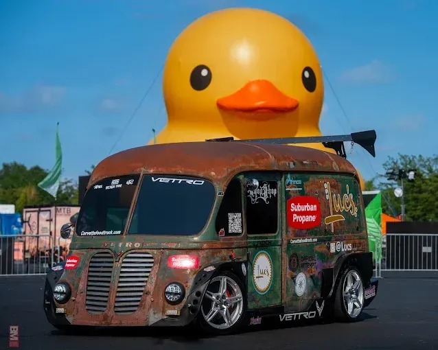 Custom brown van parked before a giant yellow duck sculpture under a blue sky