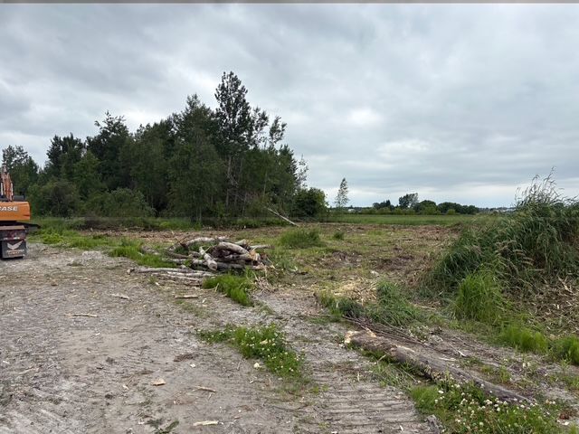 Chemin de gravier longeant un champ boueux et des arbres sous un ciel nuageux, avec une pelleteuse partiellement visible à gauche.