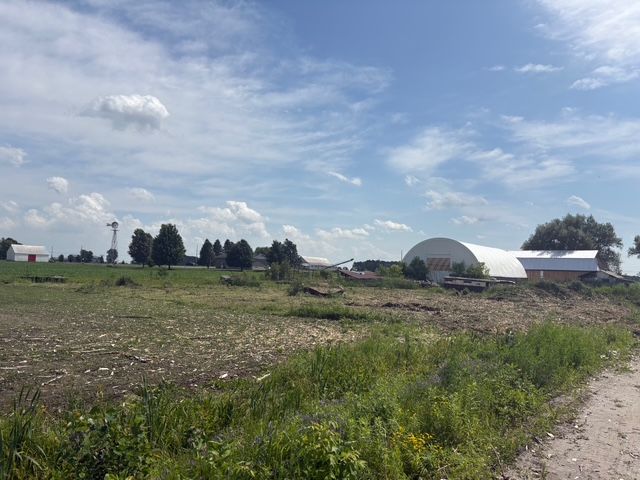 Champ agricole sous un ciel bleu nuageux, avec des granges et une serre en forme de dôme au loin.