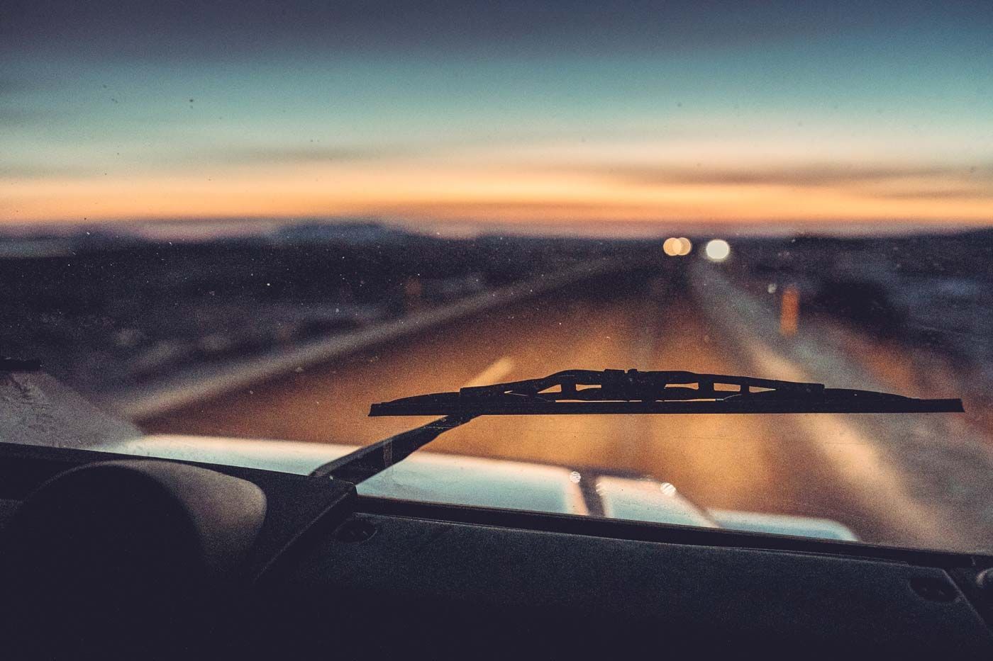 Vista do painel de um carro ao entardecer, olhando para uma estrada em direção aos faróis ao longe.