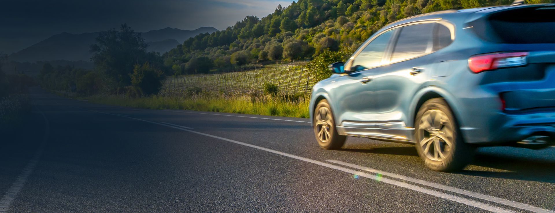 Blue car driving on a sunlit mountain road beside green hills | Coastal Automotive Repair