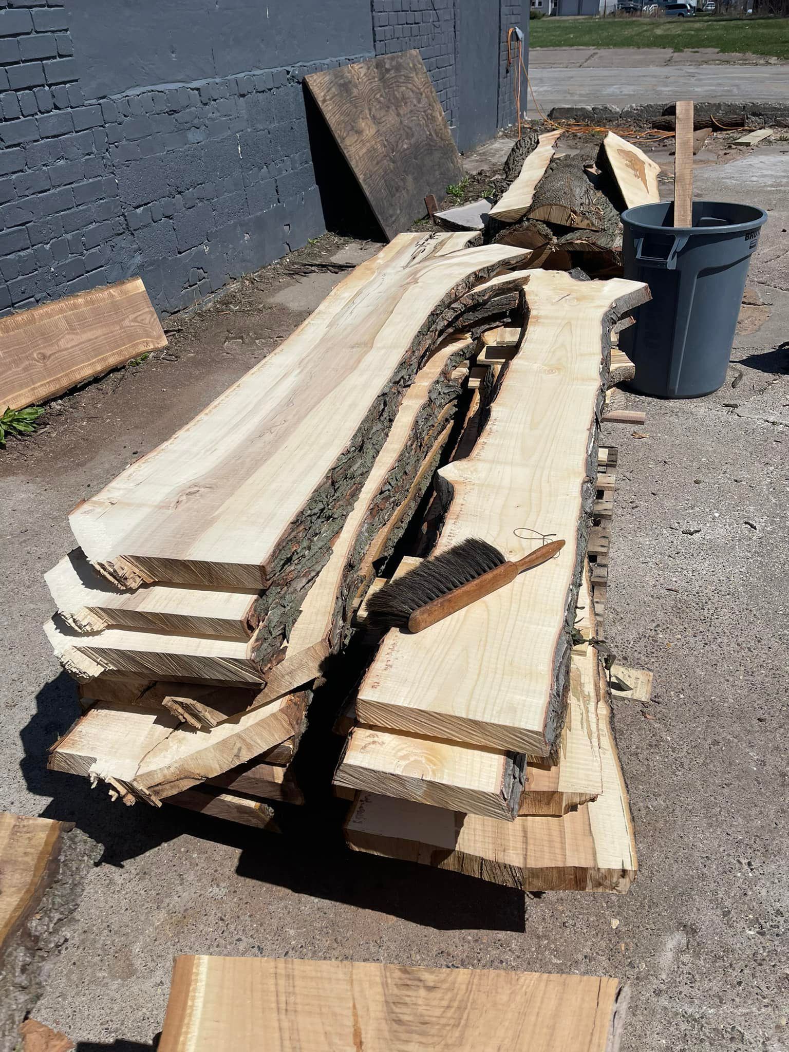 Stacked rough-cut lumber boards on gravel beside a building wall and trash bin outdoors.